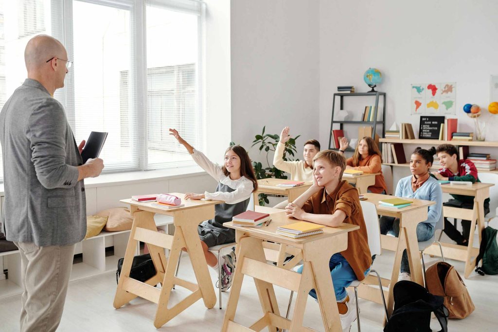 A teacher standing in front of a Middle School classroom. One student raises their hand.